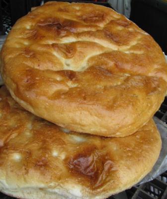 Dwa okrągłe, złociste chleby Pide ułożone jeden na drugim, widoczne delikatne wgłębienia na powierzchni, sugerujące turecki styl, na tle ciemnej kratki.